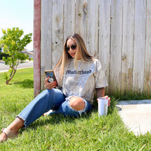 Load image into Gallery viewer, Woman sitting on grass wearing a &#39;Motherhood&#39; shirt with a wooden fence and trees in the background.