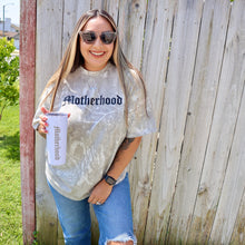 Load image into Gallery viewer, Woman wearing a &#39;Motherhood&#39; shirt holding a baby bottle outdoors.