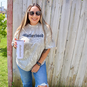 Woman wearing a 'Motherhood' shirt holding a baby bottle outdoors.